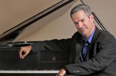 Pianist Robert Thies sits in front of a Steinway grand piano. The lid is open and the keys are uncovered. Thies sits with his back to it, his arm resting on the lifted fallboard.