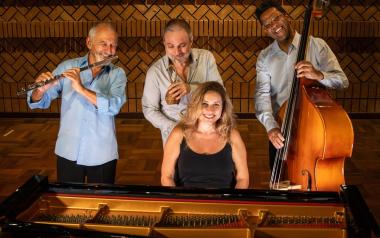 The Heloísa Fernandes Quartet stands smiling in front of a piano, each holding their instruments 